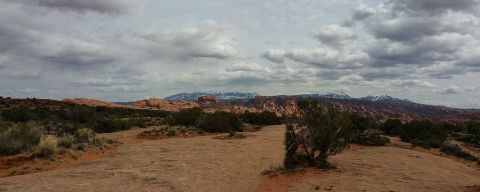 1979 Ford Bronco - A panoramic view of a desert landscape with red rock formations and snow-capped mountains in the distance under a cloudy sky.