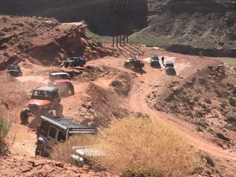 1979 Ford Bronco - A convoy of off-road vehicles, including Jeeps and a Bronco, navigates a dusty, winding dirt trail through a rocky, arid canyon landscape.