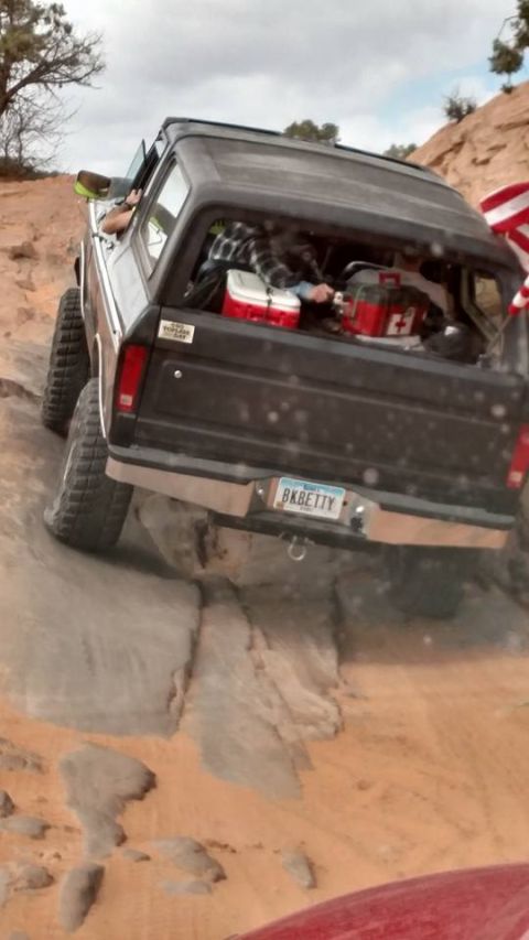 1979 Ford Bronco - The rear and side of a black 1979 Ford Bronco is shown driving off-road on a rocky, sandy incline. The vehicle has large, knobby tires and…