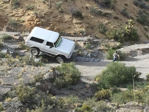 1979 Ford Bronco - A white 1979 Ford Bronco is navigating a rocky, off-road trail. A person is visible walking on the trail behind the vehicle.