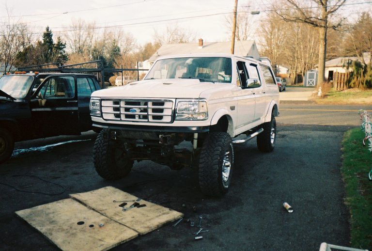 White lifted Ford SUV, front three-quarter, bumper removed, in driveway with parts on ground