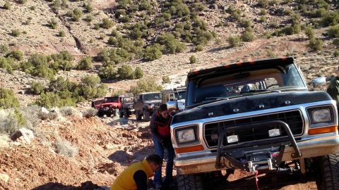 1979 Ford Bronco - A line of off-road vehicles, including a black Ford Bronco in the foreground, navigate a rocky, arid terrain.