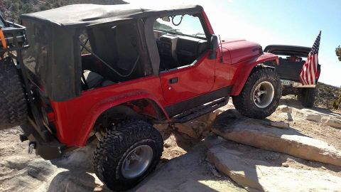 1979 Ford Bronco - A red Jeep Wrangler is navigating a rocky trail, with its front tire perched on a large rock. A black pickup truck with an American flag is…