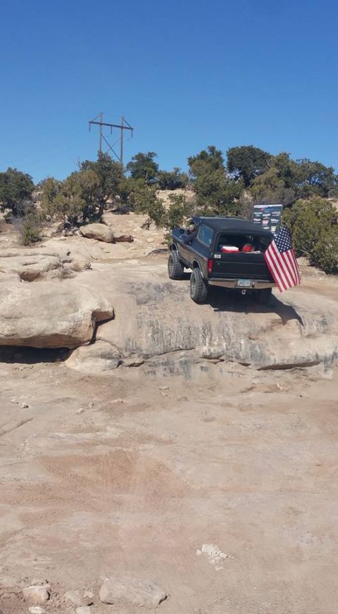 1979 Ford Bronco - A black Ford Bronco is parked on a rocky, desert-like terrain under a clear blue sky. An American flag is attached to the rear of the vehic…