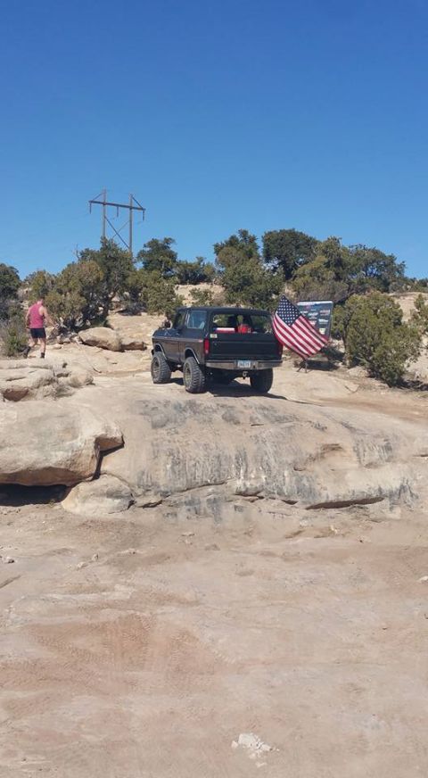 1979 Ford Bronco - A black 1979 Ford Bronco is parked on a rocky, desert trail with an American flag displayed on the side. A person in a red shirt and shorts…