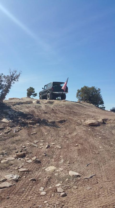 1979 Ford Bronco - A black SUV with an American flag attached to the rear is parked on a dirt hill under a clear blue sky.