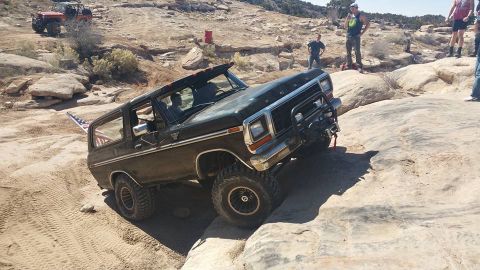 1979 Ford Bronco - A dark-colored Ford Bronco is navigating a rocky, off-road trail, tilted at a steep angle. An American flag is visible in the rear passenge…