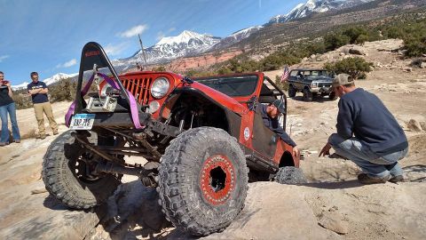 1979 Ford Bronco - A modified red Jeep is navigating a rocky trail with two people observing. In the background, a dark Ford Bronco is parked.