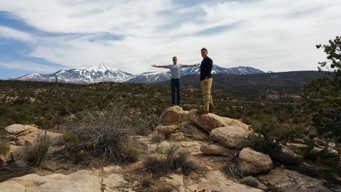1979 Ford Bronco - Two men stand on rocks in a rocky, arid landscape with snow-capped mountains in the background under a cloudy sky.