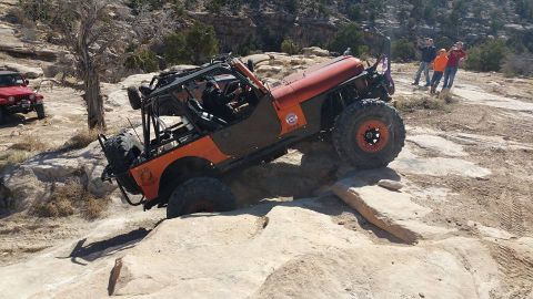1979 Ford Bronco - An orange Jeep Wrangler is navigating a rocky, off-road trail. The vehicle is tilted at an angle as it climbs over large rocks, with a driv…