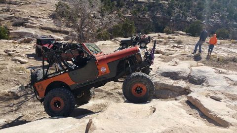 1979 Ford Bronco - An orange and olive green modified Jeep is navigating a rocky, off-road trail. Another red Jeep is visible behind it, and two people stand…