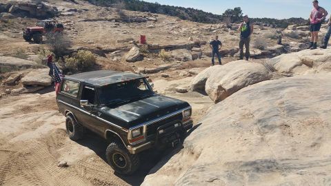 1979 Ford Bronco - A black 1979 Ford Bronco is parked on a rocky, off-road trail with several people standing around. An American flag is attached to the rear…