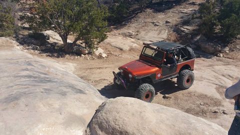 1979 Ford Bronco - An orange Jeep CJ is navigating a rocky, off-road trail with a driver visible behind the wheel. The vehicle has large off-road tires and a…