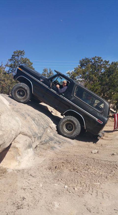 1979 Ford Bronco - A black 1979 Ford Bronco is shown off-roading on a rocky incline under a clear blue sky. The driver is visible behind the wheel, and the ve…