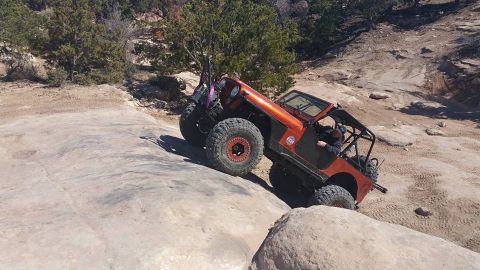 1979 Ford Bronco - An orange Jeep Wrangler is navigating a steep, rocky incline on an off-road trail. The vehicle has large, knobby tires and a roll cage, ind…