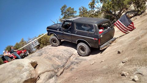 1979 Ford Bronco - A black 1979 Ford Bronco is shown on a rocky incline with an American flag attached to the rear. Another red Jeep is visible in the backgro…
