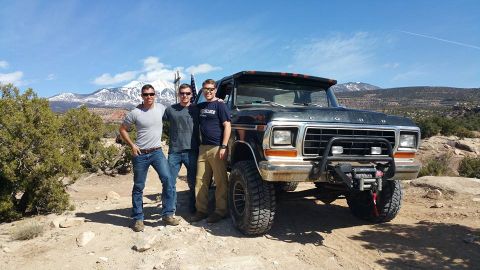 1979 Ford Bronco - Three men pose in front of a lifted, black 1970s Ford truck with large tires and a winch on the front bumper. The truck is parked on a dirt…