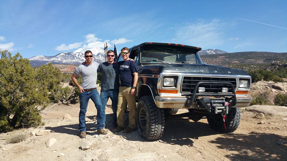 Three men stand next to a lifted black Ford truck with a winch and large tires, mountains in the background.