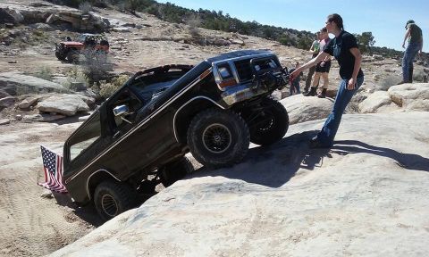 1979 Ford Bronco - A black 1979 Ford Bronco is navigating a rocky incline with an American flag attached to the rear. Several people are observing the vehicle…