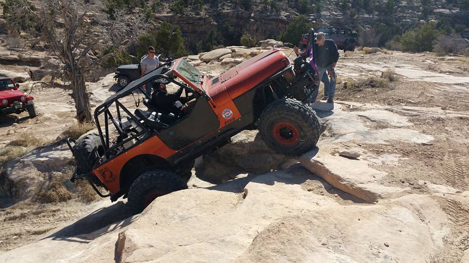 Orange Jeep Wrangler with roll cage climbing over rocks, red Jeep in background.