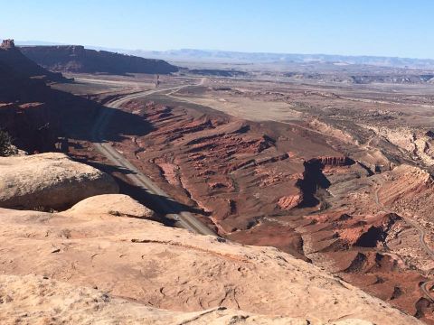 1979 Ford Bronco - A vast desert landscape with red rock formations and a winding road cutting through a canyon under a clear blue sky.