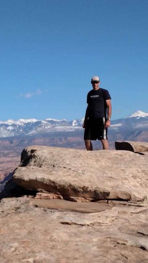 1979 Ford Bronco - A man wearing a black t-shirt and shorts stands on a rocky outcropping with snow-capped mountains in the background.
