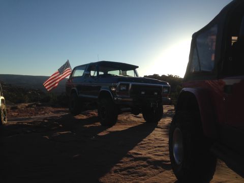 1979 Ford Bronco - A dark blue Ford Bronco is parked on a rocky surface with an American flag attached to its side. The sun is setting behind the vehicle, cas…