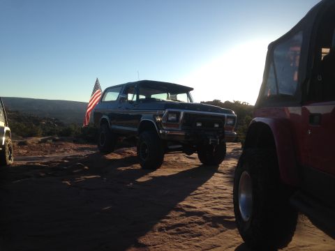 1979 Ford Bronco - A dark blue 1979 Ford Bronco with an American flag attached to the rear is parked on a rocky, desert landscape under a clear sky.