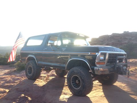 1979 Ford Bronco - A dark colored Ford Bronco is parked on a rocky, desert terrain with an American flag attached to the rear passenger side. The sun is low i…