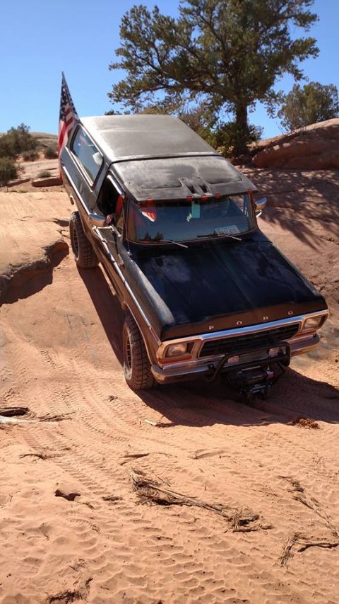 1979 Ford Bronco - A black Ford Bronco with an American flag attached to the rear is navigating a sandy, off-road trail with tire tracks visible in the dirt.…