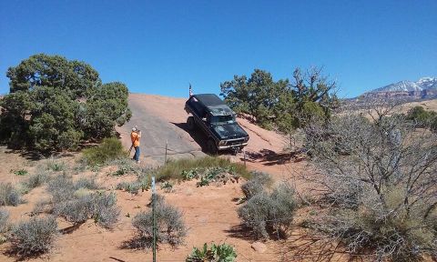1979 Ford Bronco - A black Ford Bronco is driving up a steep, rocky incline in a desert landscape. A person in an orange shirt is standing to the left, holdin…