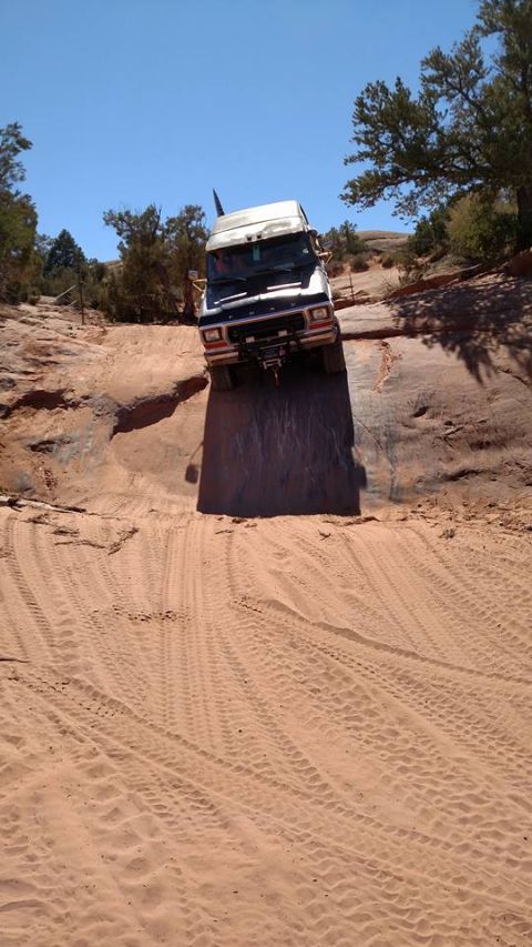 1979 Ford Bronco - A dark-colored van is driving down a steep, sandy incline in a desert landscape with sparse trees and a clear blue sky.