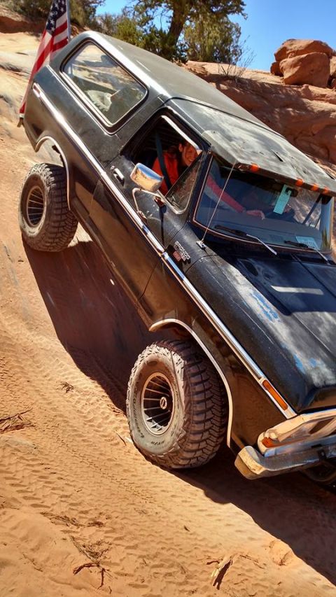 1979 Ford Bronco - A black 1979 Ford Bronco is shown off-roading on a steep, sandy incline with large, aggressive tires. An American flag is mounted on the re…