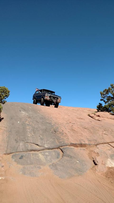 1979 Ford Bronco - A dark colored SUV is driving up a steep, rocky incline under a clear blue sky. An American flag is visible on the roof rack of the vehicle.