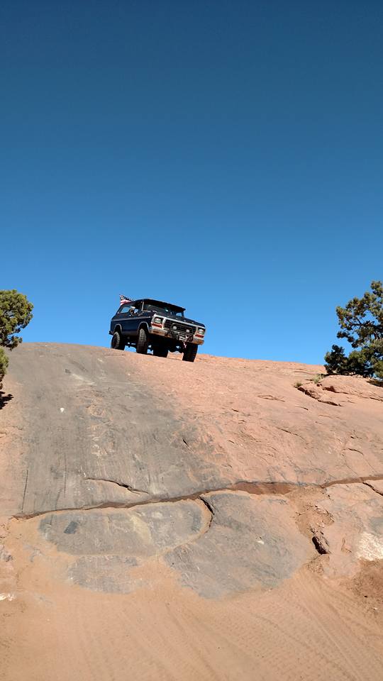 Dark colored SUV driving up a rocky hill with an American flag on the roof.