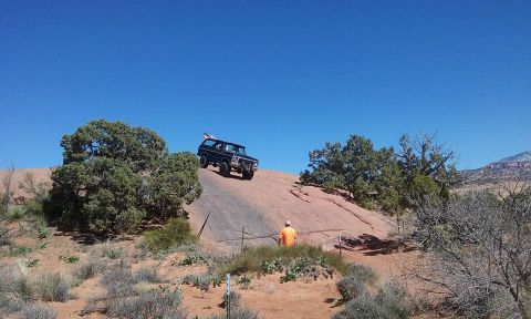 1979 Ford Bronco - A dark colored Ford Bronco is driving up a steep, rocky incline under a clear blue sky. A person wearing an orange shirt stands on the side…