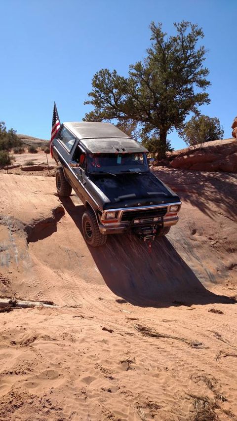 1979 Ford Bronco - A black, lifted 1979 Ford Bronco is shown off-roading on a sandy, rocky incline under a clear blue sky. An American flag is mounted on the…