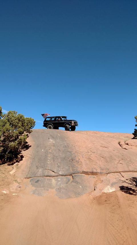 1979 Ford Bronco - A black Ford Bronco with an American flag attached to its rear is parked on a rocky, sandy incline under a clear blue sky.