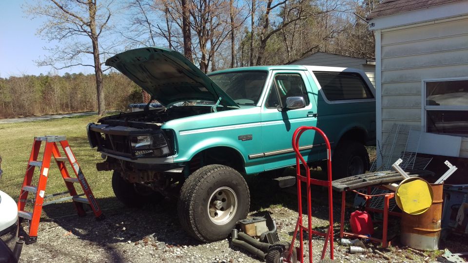Teal 1994 Ford Bronco with hood open, parked next to a building with an orange ladder.