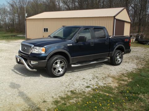 2005 Ford F-150 - A dark blue Ford F-150 Supercrew pickup truck is parked on a gravel lot in front of a tan metal building. The truck has a chrome bull bar o…