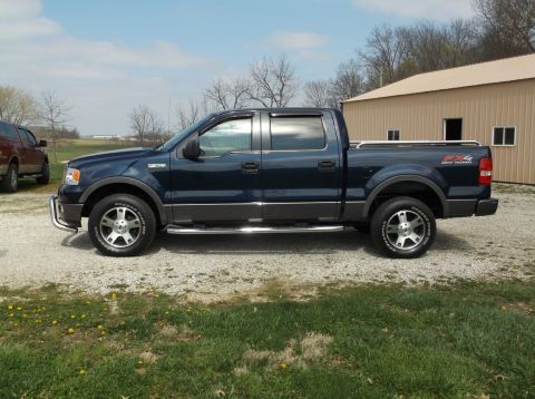 2005 Ford F-150 - A dark blue Ford F-150 Supercrew pickup truck is parked on a gravel lot next to a tan building. The truck is shown from the side view, feat…