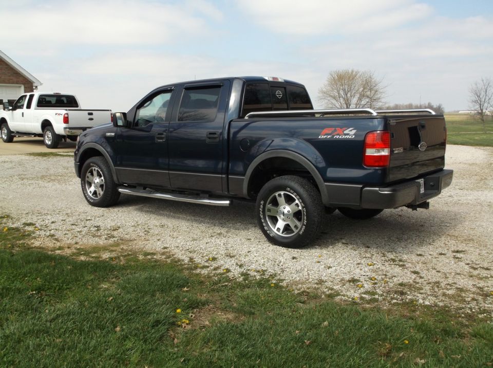 Dark blue Ford F-150 pickup truck parked on gravel, white Ford F-150 behind it.