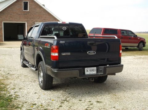 2005 Ford F-150 - The rear driver's side of a dark blue Ford F-150 pickup truck is shown parked on a gravel driveway in front of a brick building. A red crew…