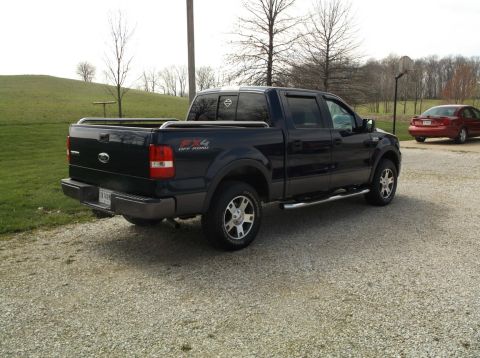 2005 Ford F-150 - A dark blue Ford F-150 pickup truck is parked on a gravel surface with a red sedan in the background.