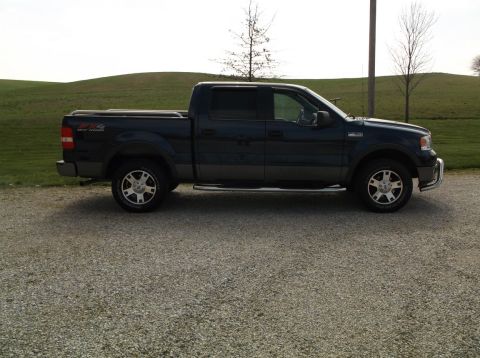 2005 Ford F-150 - A dark blue pickup truck is parked on a gravel surface with a grassy hill in the background.