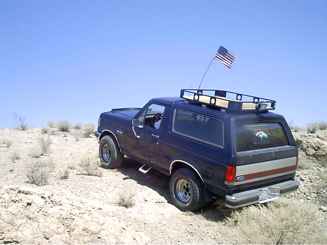 Dark blue Ford Bronco with roof rack and American flag parked on a desert hillside, rear three-quarter view