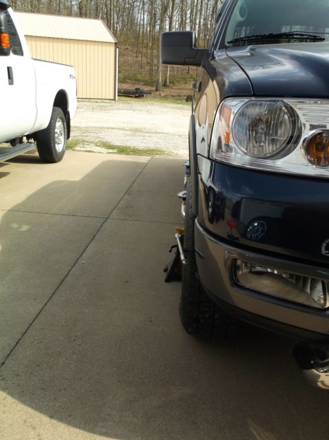 2005 Ford F-150 - A dark blue Ford F-150's front wheel is elevated on a jack stand, with a white truck visible to the left.