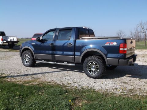 2005 Ford F-150 - A blue pickup truck is parked on a gravel lot under a clear blue sky. A white truck is visible in the background.