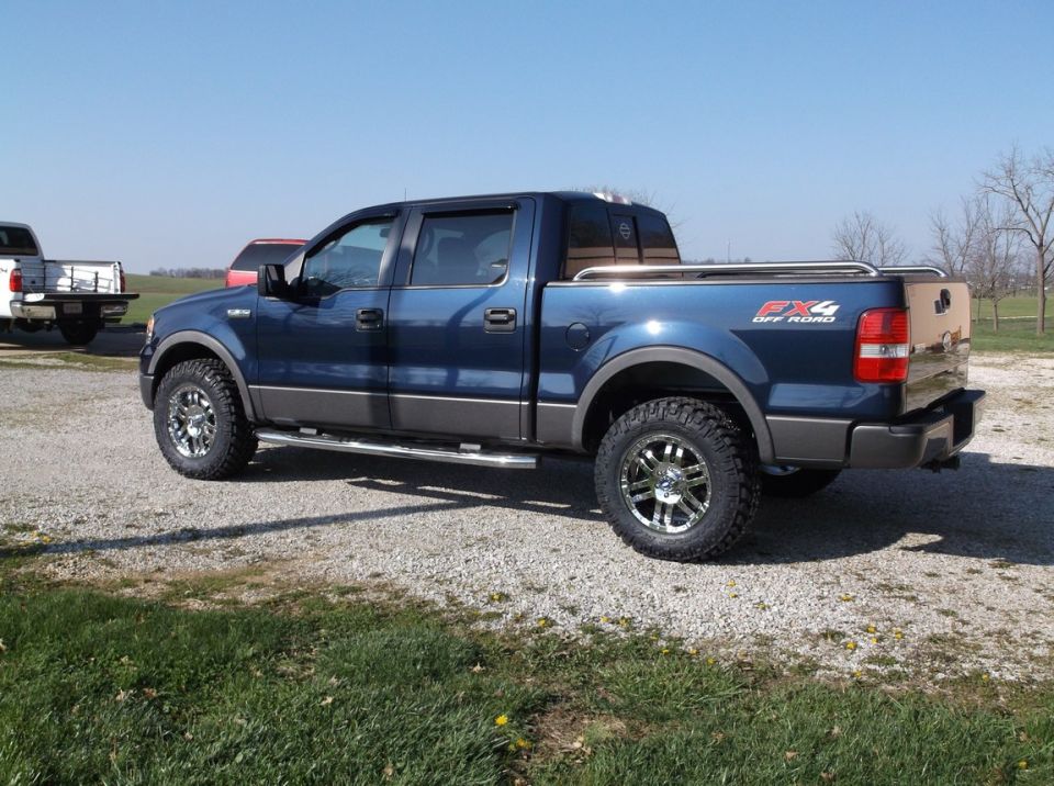 Blue pickup truck with chrome wheels and off-road tires. Another white truck is parked behind it