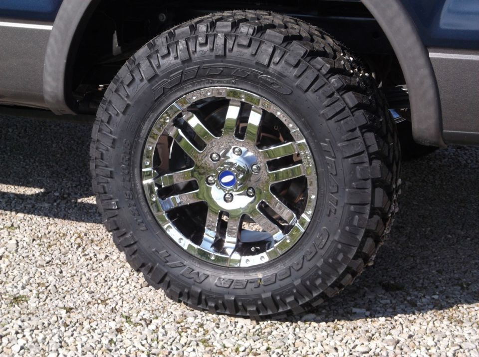 Chrome alloy wheel with a blue center cap on a large off-road tire. Part of a dark-colored truck is visible